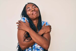 African american woman with braids wearing casual summer clothes hugging oneself happy and positive, smiling confident. self love and self care